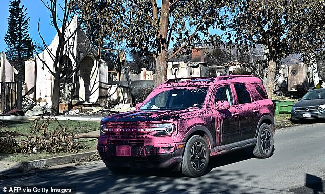 A press vehicle covered in pink flame retardant drives past homes destroyed in the Palisades Fire in Pacific Palisades