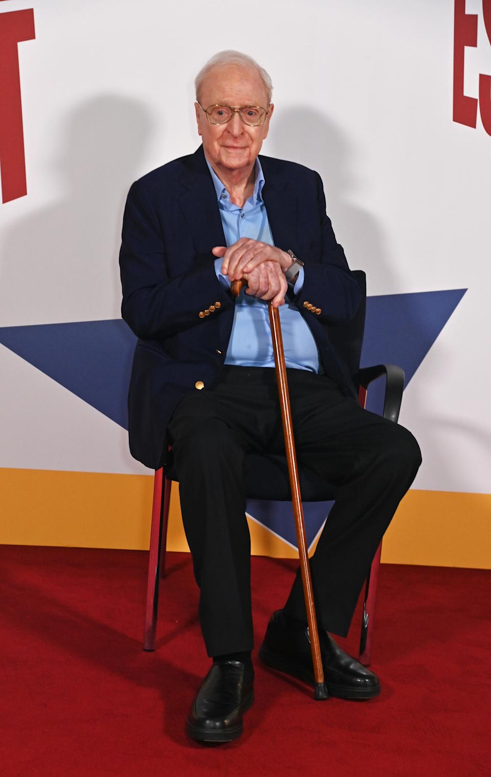 Elderly man in a suit seated on a red carpet, holding a cane, looking at the camera at a celebrity event