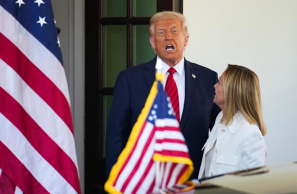 A man in a suit and red tie is speaking loudly beside a woman in a white coat, near American flags