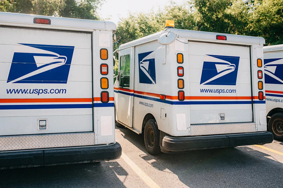 Three USPS delivery trucks are parked, showcasing the postal service logos and website addresses.