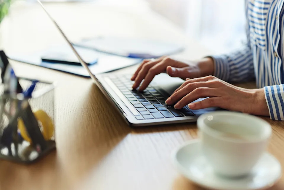 Person typing on a laptop at a wooden desk with a coffee cup nearby, wearing a striped shirt.