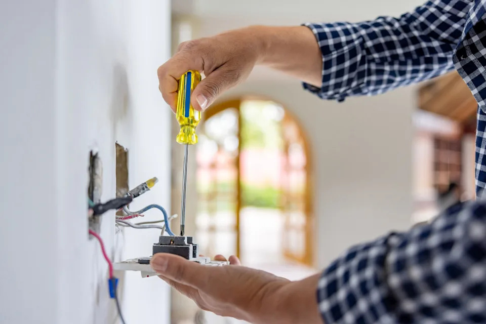 Person installing a light switch with a screwdriver, wearing a checkered shirt.