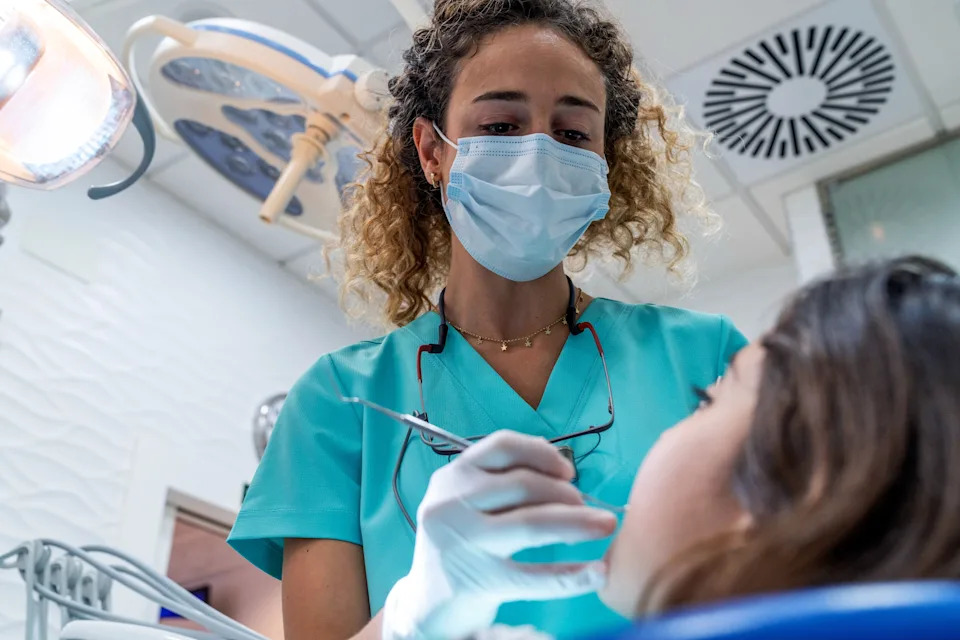 A dental employee wearing a mask and scrubs examines a patient in a dentist's office.