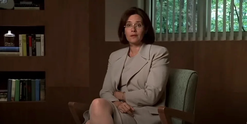 A person sits in an office setting, dressed in a formal suit, with shelves and a window in the background