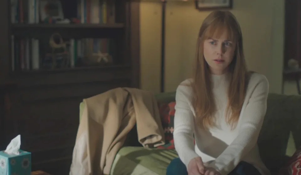 Person with long hair sits on a couch, wearing a light sweater, looking contemplative. A coat is draped over the couch