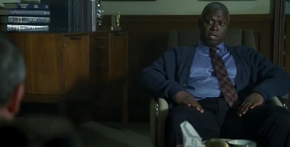 A man in an office setting sits in a chair, wearing a cardigan and tie, appearing engaged in a conversation