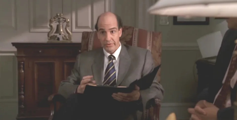 A man in a suit and tie holds a folder, sitting in an office chair, appearing to be speaking or listening intently