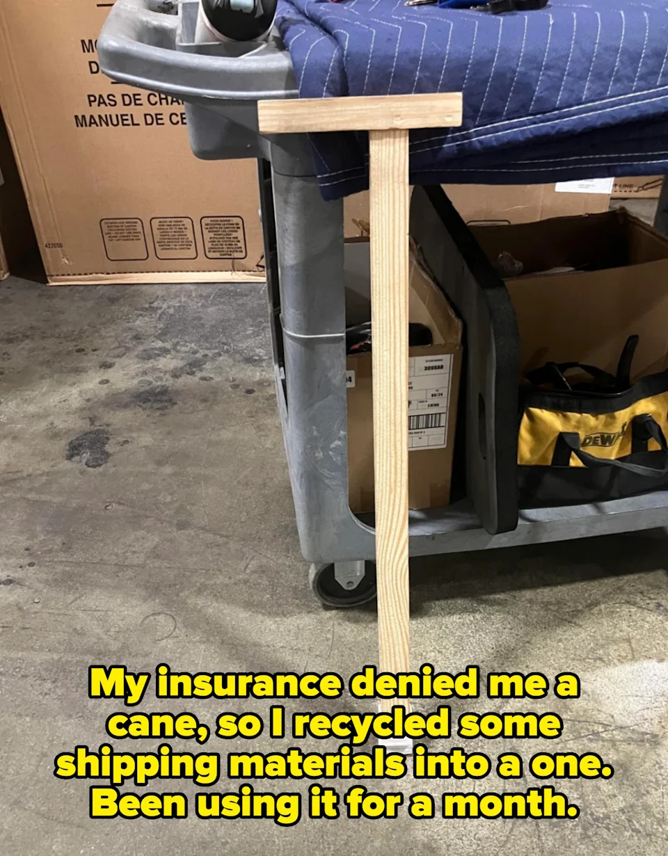 Wooden tool with a T-shape handle leaning against a cart in a warehouse setting. Caption: "My insurance denied me a cane, so I recycled some shipping materials into a one. Been using it for a month"