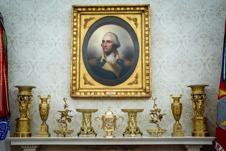 A gilded portrait of George Washington hangs above ornate gold decorations on a mantle, with ceremonial flags in the background