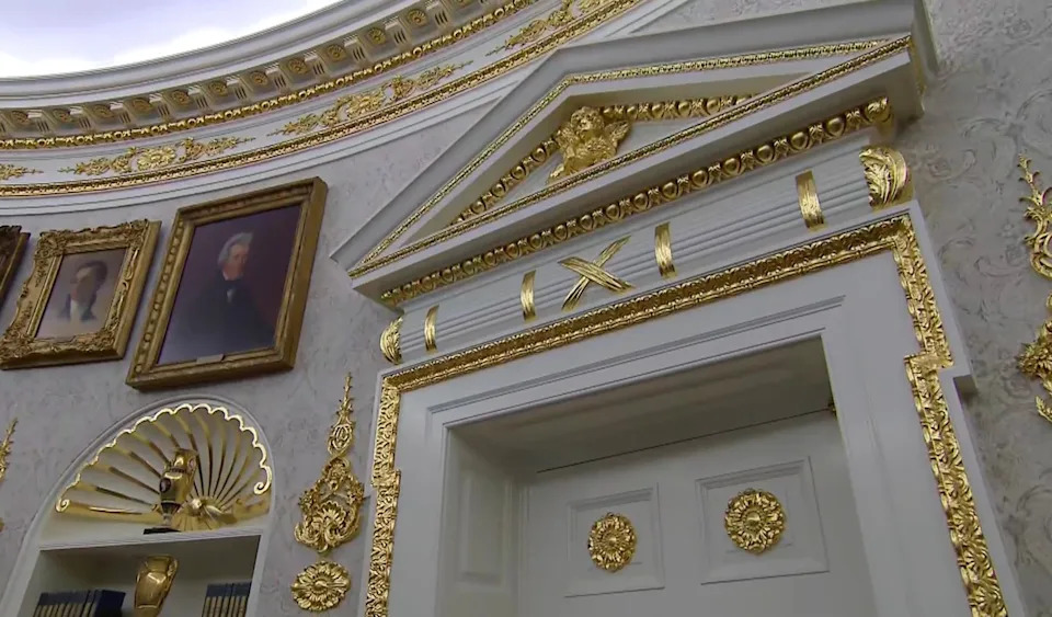 Ornate architectural details with gold accents and framed portraits decorate an elegant, historically styled room
