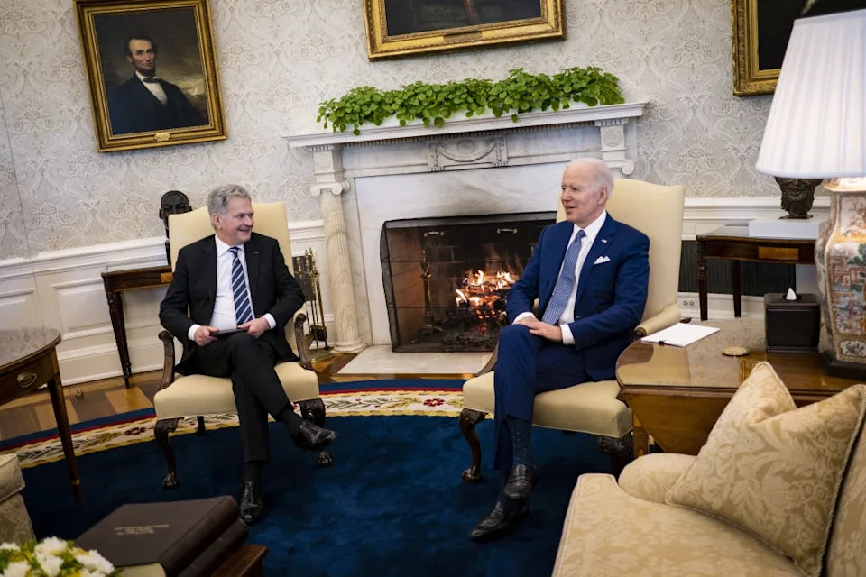 Two men in formal suits are seated in an elegant room with a fireplace, having a conversation