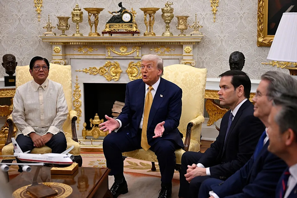 Several people in formal attire are seated and listening attentively in an ornate room with a model airplane and decorative elements