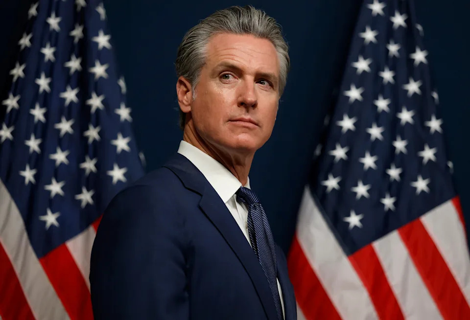 A person in a suit stands in front of two U.S. flags, looking to the side with a serious expression