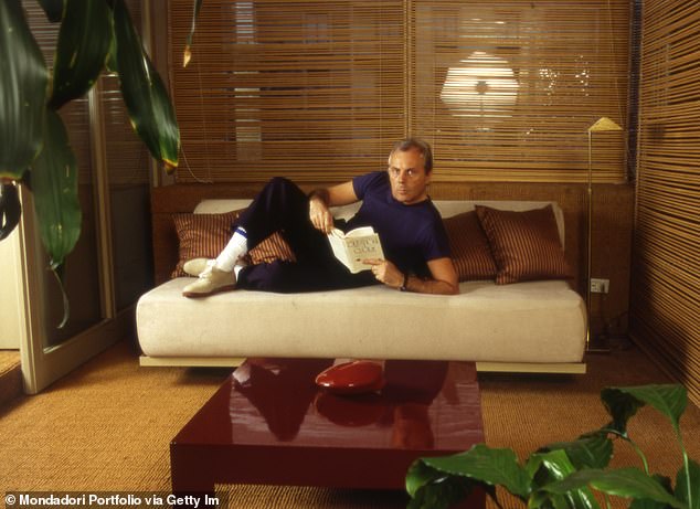 The Italian fashion stylist poses with a book on the sofa in his living room in Italy in 1984