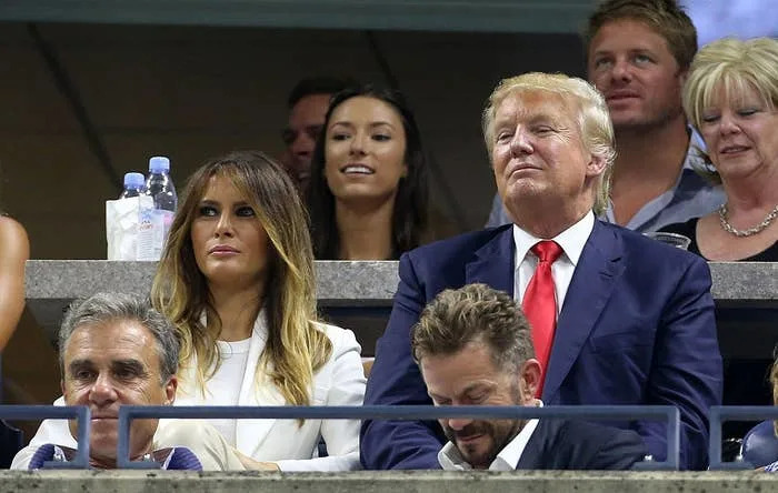 A group of people in stadium seats, one man in a suit and tie, and a woman in a stylish outfit. Others are seated around them