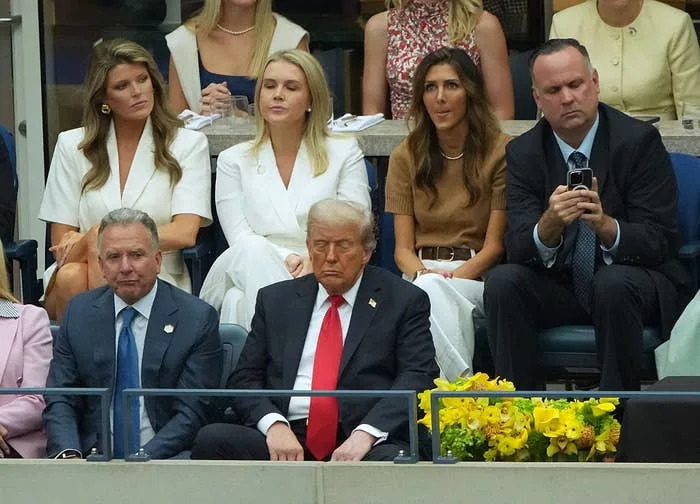 A group of people seated at an event. A man in a suit with a red tie sits in the front row, appearing serious. Other attendees are behind him