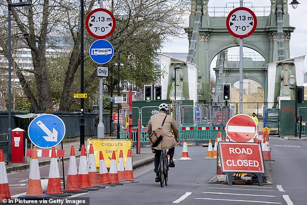 A local MP recently warned the current repair schedule for Hammersmith may take another ten years to complete. Pictured: A cyclist cycles between the cones and barriers which block the southern end of Hammersmith Bridge in April 2022