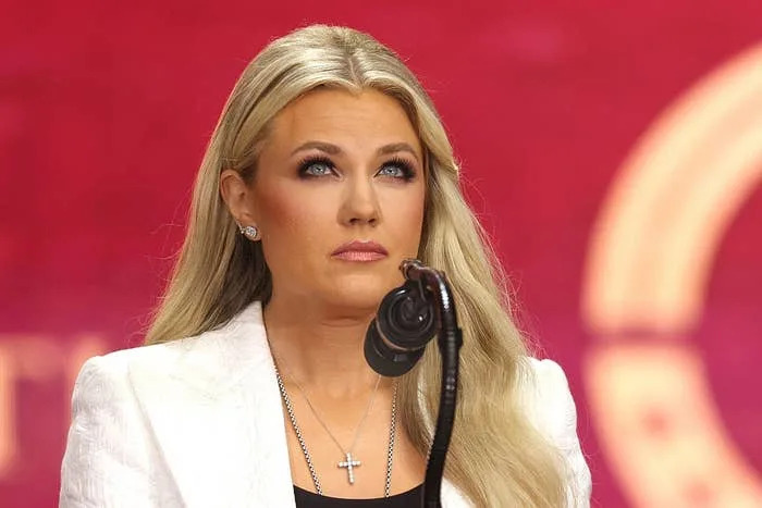 Person speaking at an event, wearing a white blazer and cross necklace, with a focused expression