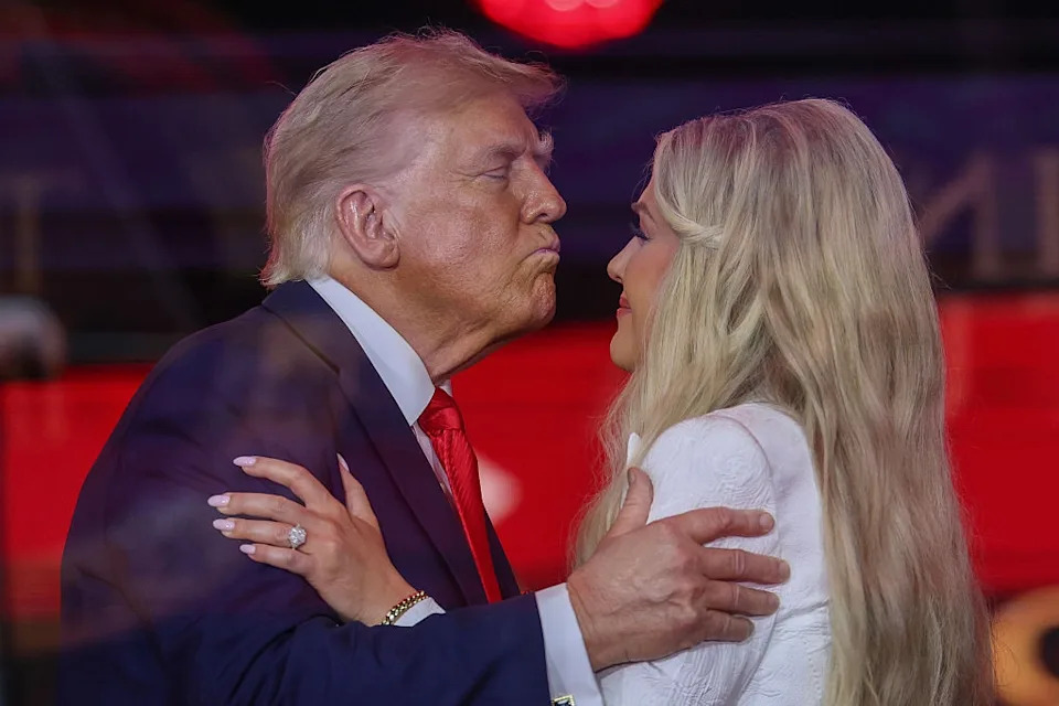 Two people embrace and lean in as if to kiss at an event. One person is wearing a suit, and the other is in a patterned outfit