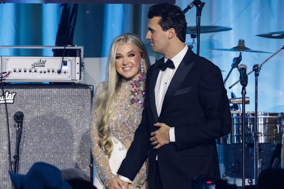 A couple on stage; the woman in a sequined dress with floral accents, the man in a tuxedo. They are smiling and holding hands