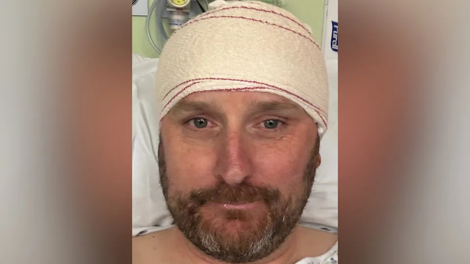 Mr Whitaker has a beard and is resting in a hospital bed, wearing a gown and a head bandage following a cranial surgery or treatment. Medical equipment is visible in the background.