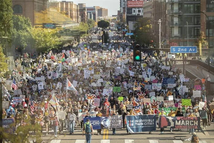 Large crowd marching in a protest, holding signs with various messages, on a city street. The event seems organized and peaceful