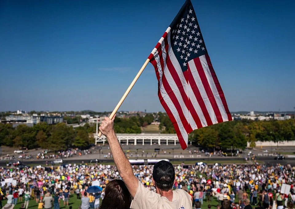 Person holding the U.S. flag overlooking a large outdoor gathering, with many people visible in the background