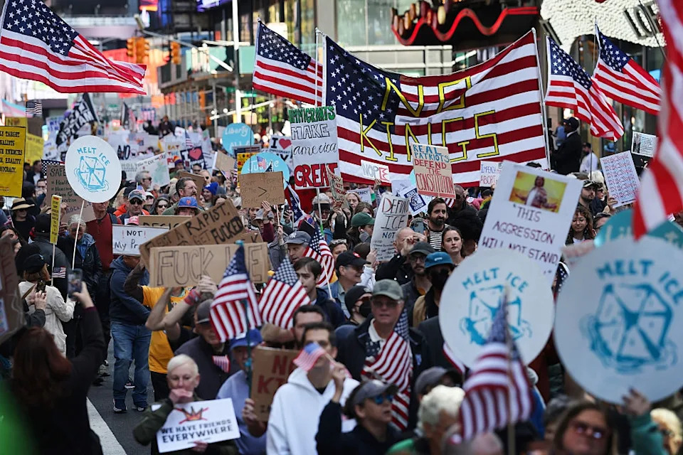 A large crowd protests on a city street, holding American flags and signs reading messages like "No KKK" and "Melt ICE."