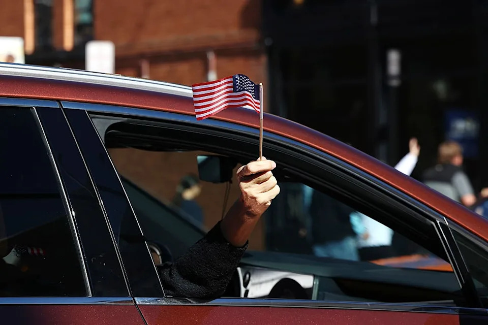 A person holds a small American flag out of a car window, participating in a parade
