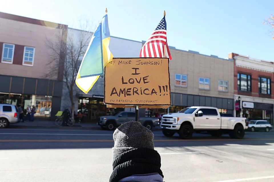 Person holding a sign reading "I Love America" with US and Ukrainian flags attached, standing on a street with buildings and vehicles in the background