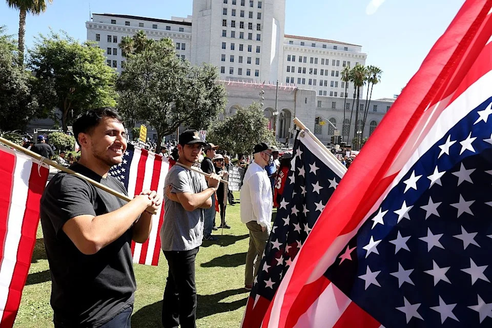 People holding US flags gather outside a large building during a daytime event