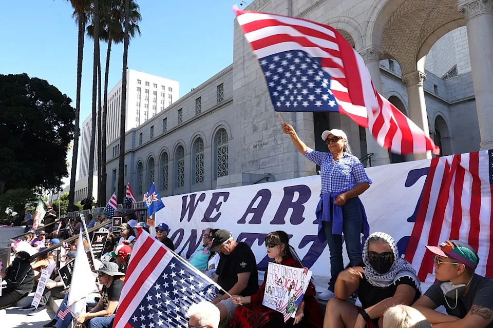 People gathered outside a building, holding American flags, with a large banner in the background. A person waves a flag, with others seated nearby
