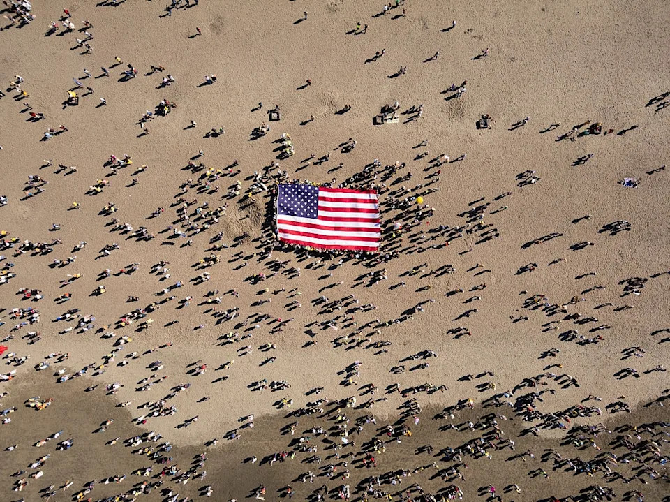 Aerial view of a crowd gathered around a large American flag on a beach, suggesting a public event or gathering