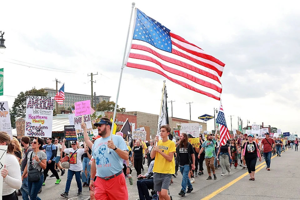 A large group of people marching peacefully, holding protest signs, with an American flag in the forefront