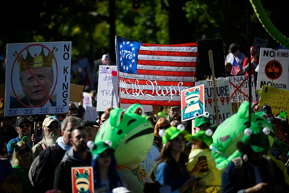 Protesters hold signs and banners, including a faux American flag and images of frogs, at a political rally