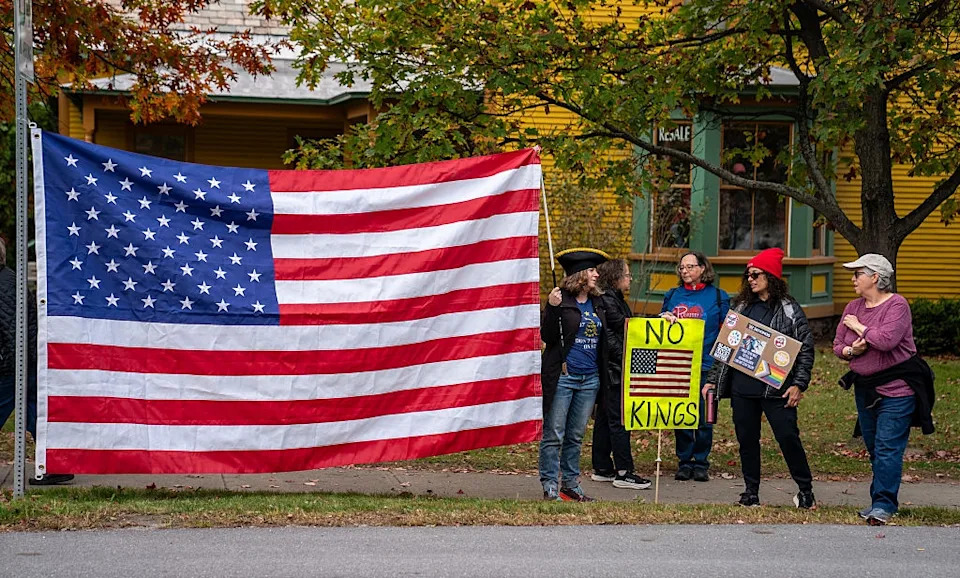 Five people protest by a large American flag. One holds a "No Kings" sign, another a framed photo. Trees and a house are in the background