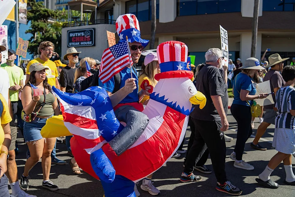 Person in an Uncle Sam-style rooster costume marches in a crowd during a protest, holding a small U.S. flag. Signs are visible in the background