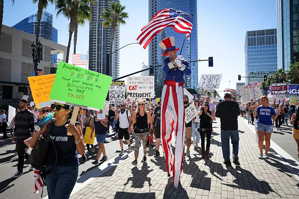 Protesters march in a city street, holding signs. A performer on stilts dressed as Uncle Sam waves an American flag amidst the crowd