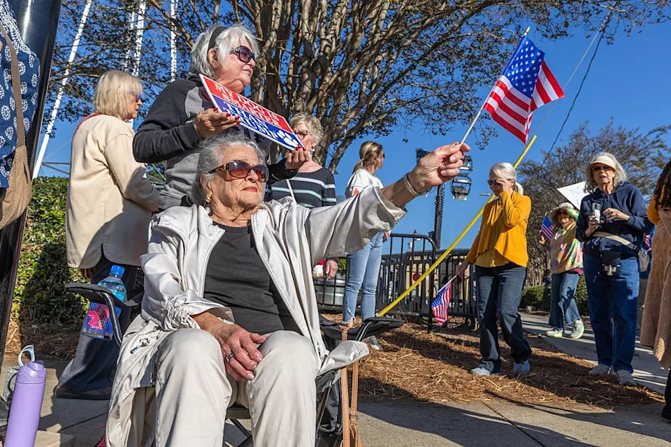 People at a rally hold American flags and signs. An older woman sits and waves a flag, engaging with the event