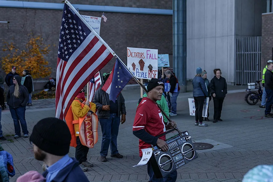 A protest scene with people holding signs. An American flag and a boombox are prominently displayed