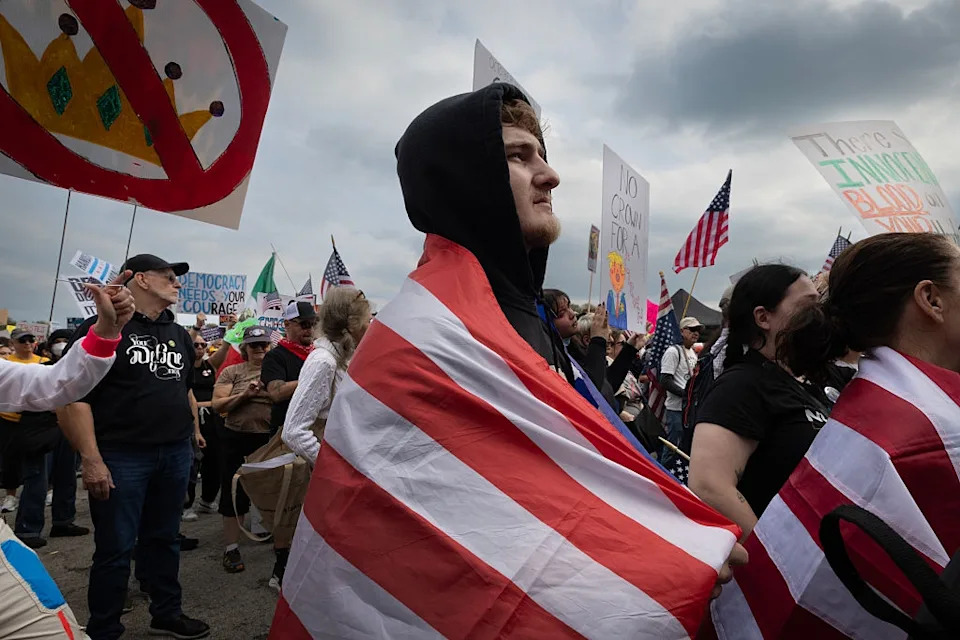 People at a protest, some holding signs and flags. A person in a hoodie draped in a striped flag is in the foreground