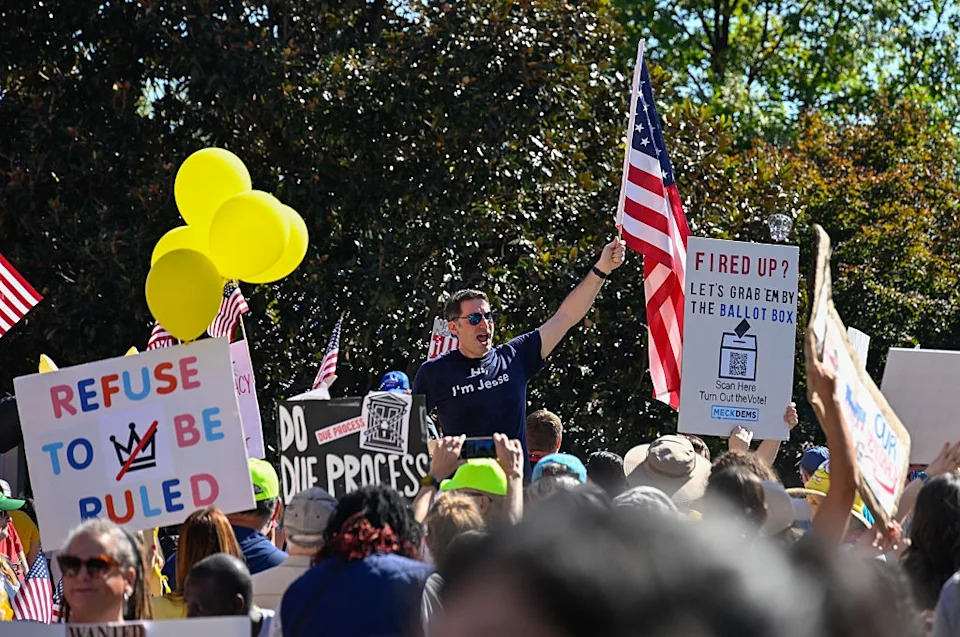 Crowd at rally holding signs like "Refuse to be ruled" and an American flag. People wear casual clothing; some have hats and sunglasses