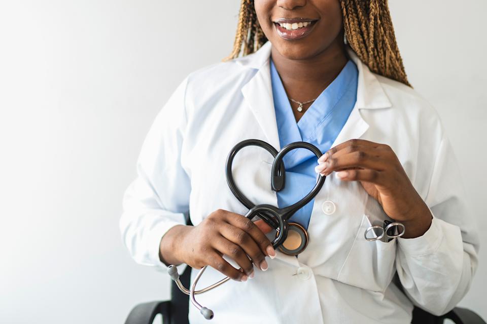 Healthcare professional with braided hair holds a stethoscope in a heart shape, smiling, wearing a white coat over scrubs