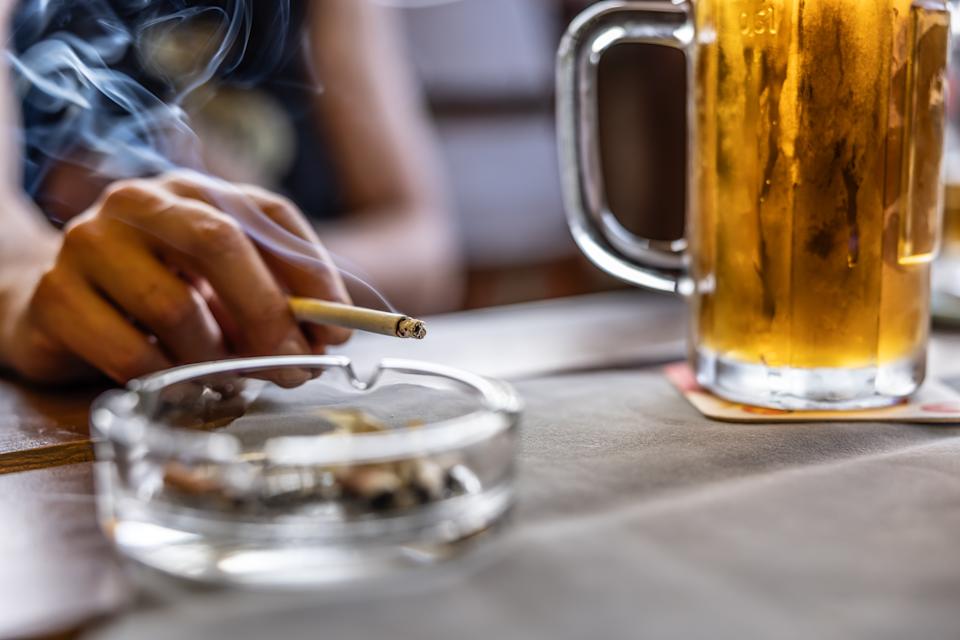 Hand holding a cigarette over an ashtray, with a glass of beer nearby on a table