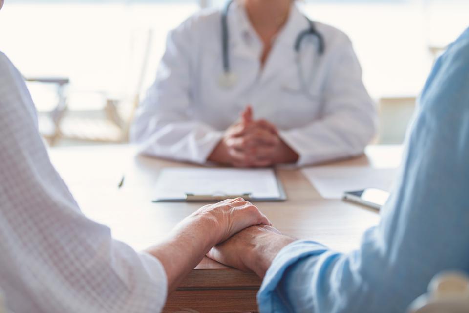 Two people holding hands at a table, facing a doctor who is speaking to them during a consultation
