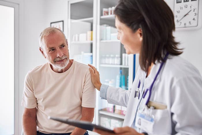 A doctor in a white coat and stethoscope comforts an older male patient seated in a medical office