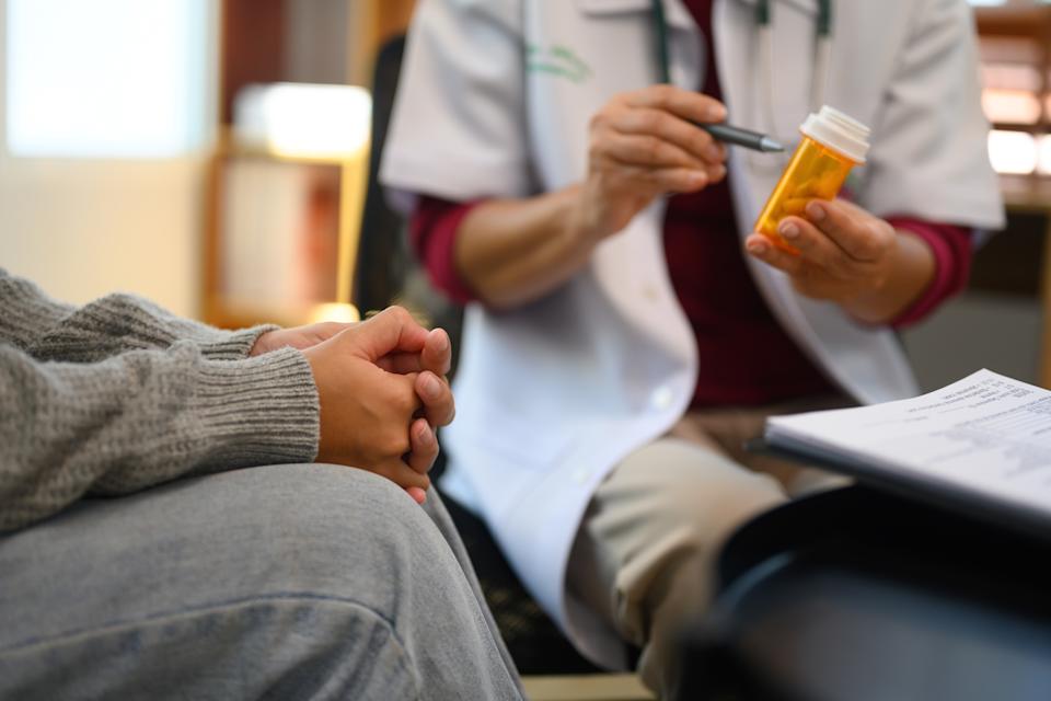 A healthcare professional discusses medication with a patient, holding a pill bottle and pointing at it, while the patient listens attentively