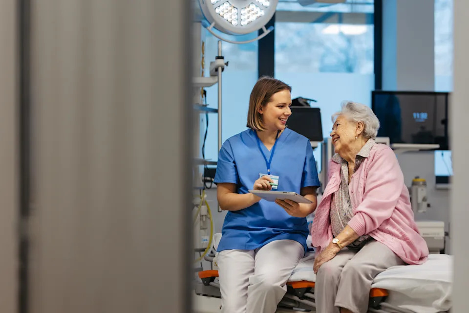 Healthcare professional talks with an elderly woman in a medical setting, both smiling, conveying a supportive interaction