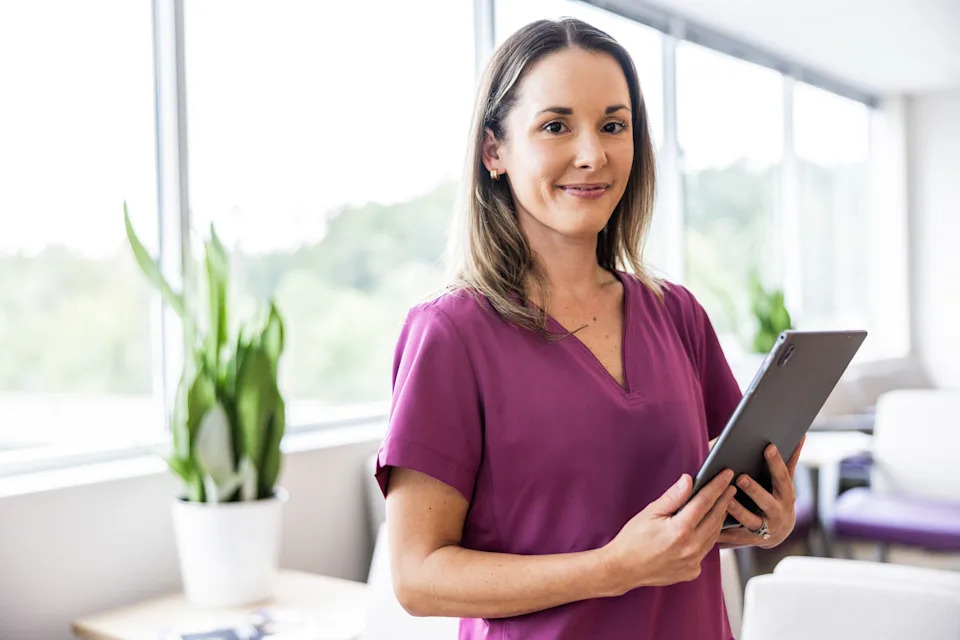 Person smiling, holding a tablet in an office with large windows and plants, conveying a professional work environment
