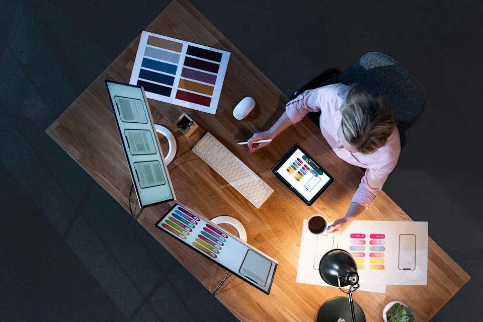 Person working at a desk with digital tablet, color swatches, and design layouts, indicating a creative design job setting. Top-down view